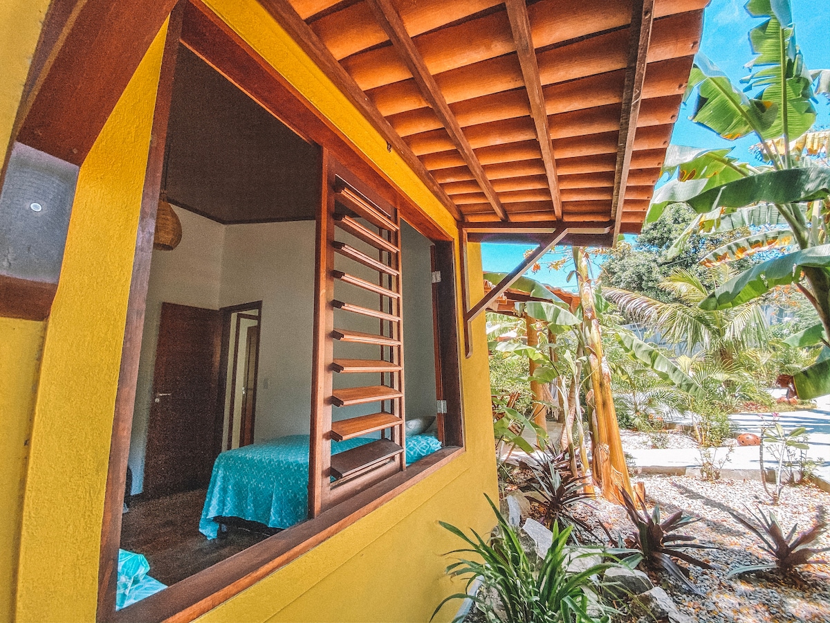 A view showcases the exterior of a brightly painted house framed by lush tropical plants. Large windows with slatted shutters offer glimpses of the interior, where a comfortable bed is partially visible. The structure is complemented by a traditional roof design, creating a welcoming ambiance.