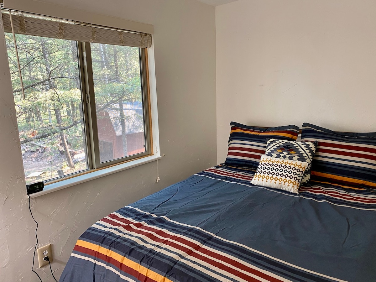 An upstairs bedroom with a queen bed and a basic layout, featuring a window with views of the surrounding trees. 