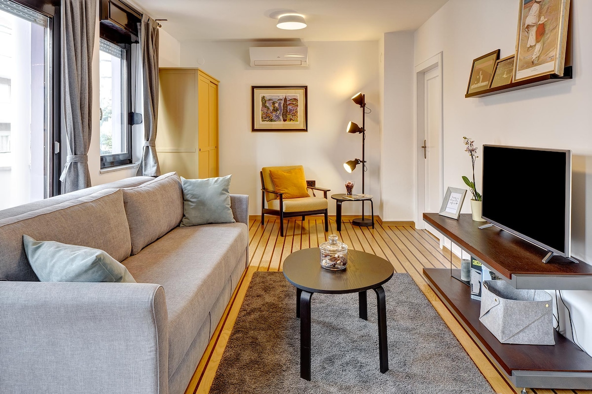 The living area features a light gray sofa with decorative cushions, a round coffee table, and a comfortable armchair. A smart TV is positioned on a wall-mounted shelf, and a floor lamp adds ambient lighting. Large windows allow natural light to fill the space.