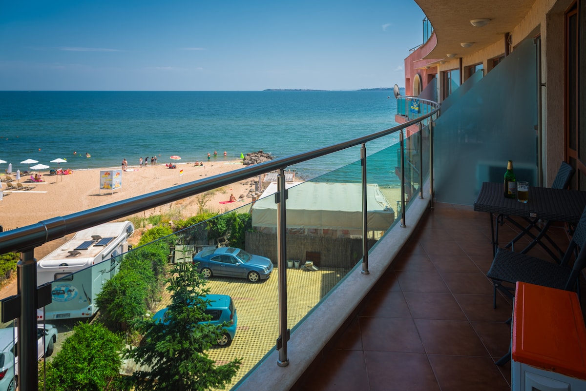A private balcony extends from the apartment, featuring a glass railing that offers unobstructed views of the sandy beach and the sea. A small table and chairs are positioned for relaxation. The distant shoreline is dotted with sun loungers and umbrellas.