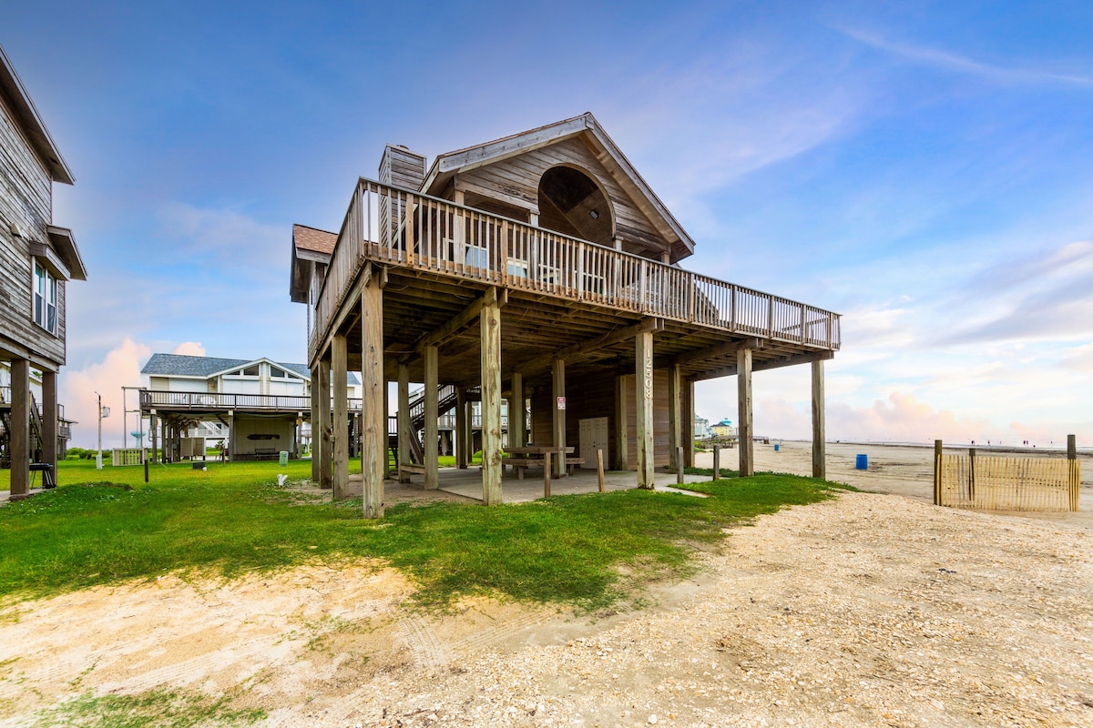 The exterior of the beach house is visible, elevated on stilts with a spacious balcony featuring wooden railings. The ground level offers an open area with picnic tables and grassy surroundings. The beach and ocean extend in the background under a clear sky, emphasizing the tranquil setting.