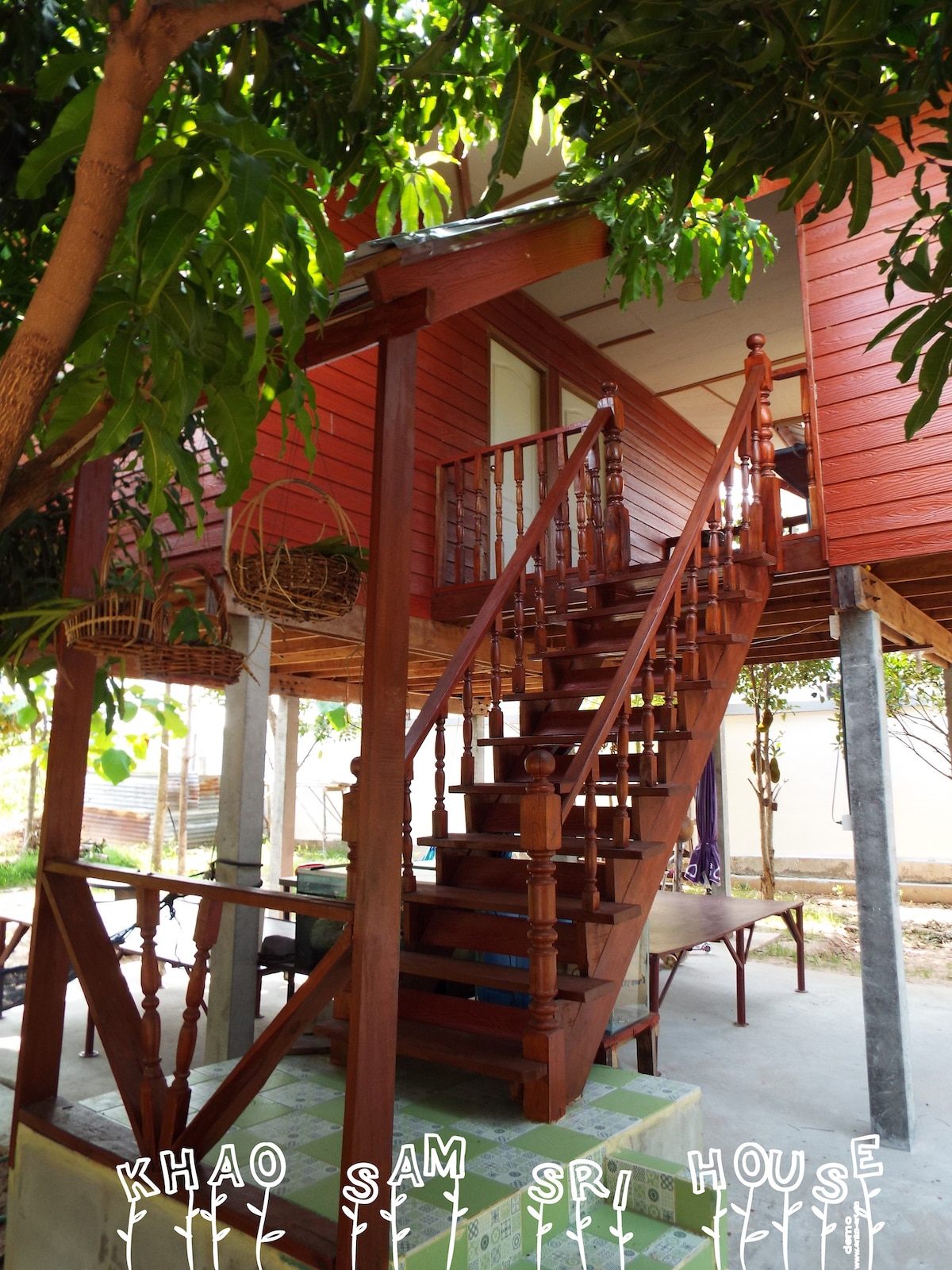 A traditional wooden house is elevated on pillars, featuring a set of sturdy stairs leading to a covered entrance. The structure is surrounded by a lush garden with greenery visible beneath the house, showcasing the natural environment of the area.
