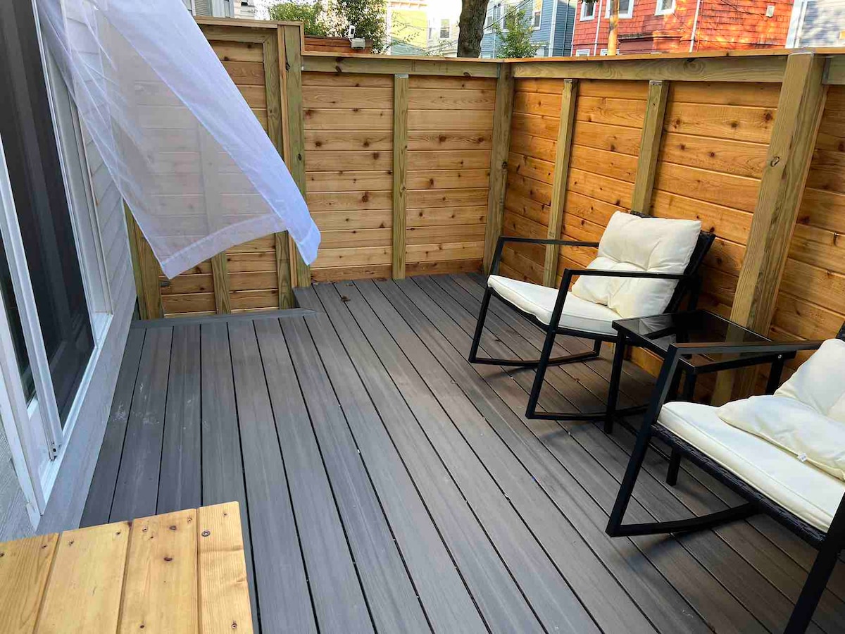 A private yard is enclosed by a wooden fence, featuring a deck with gray flooring. Two black chairs with light cushions are positioned for seating. Natural light filters through nearby sheer fabric, enhancing the inviting atmosphere of the outdoor space.
