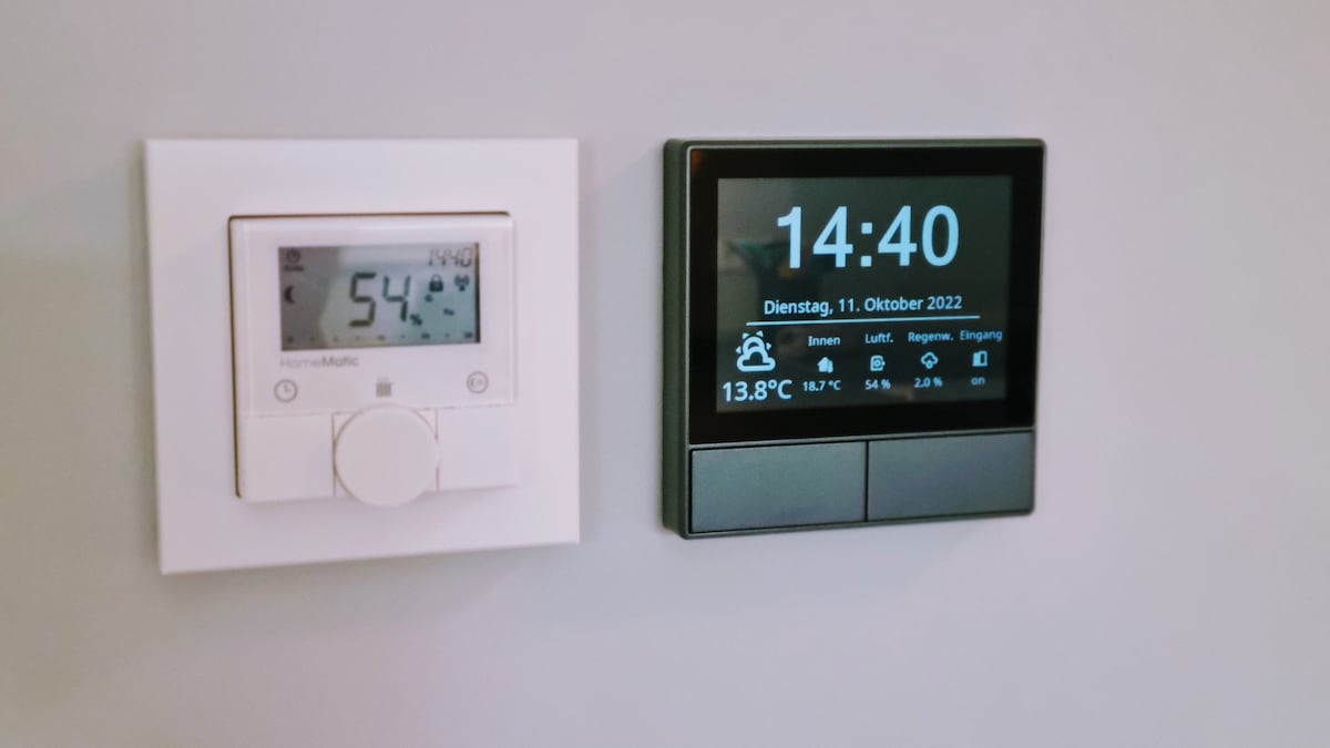 A wall is equipped with two temperature control devices. On the left, a digital thermostat displays the current humidity level and temperature. On the right, a modern touch screen shows the time and current indoor temperature, alongside additional readings.