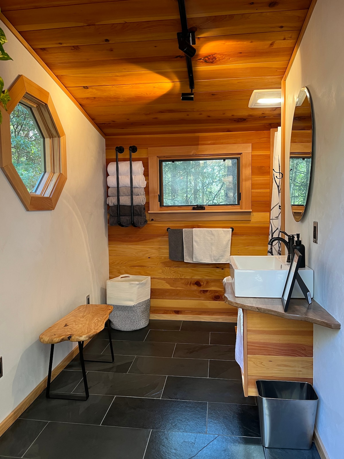 A shared bathroom area features natural wooden accents and a stone countertop with a sleek sink. A round mirror reflects warm lighting, and soft towels are neatly hung on the wall. A small wooden bench adds practicality to the space, contrasting with the dark slate flooring.