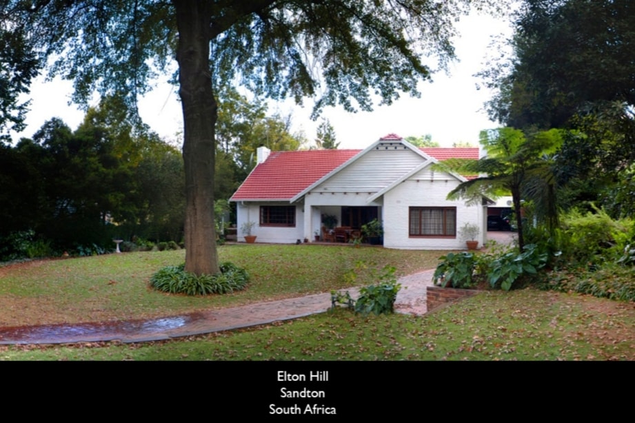 The exterior of a cottage is framed by lush greenery and trees, with a well-maintained garden leading to a paved pathway. The white structure features a red-tiled roof and large windows, suggesting ample natural light inside.