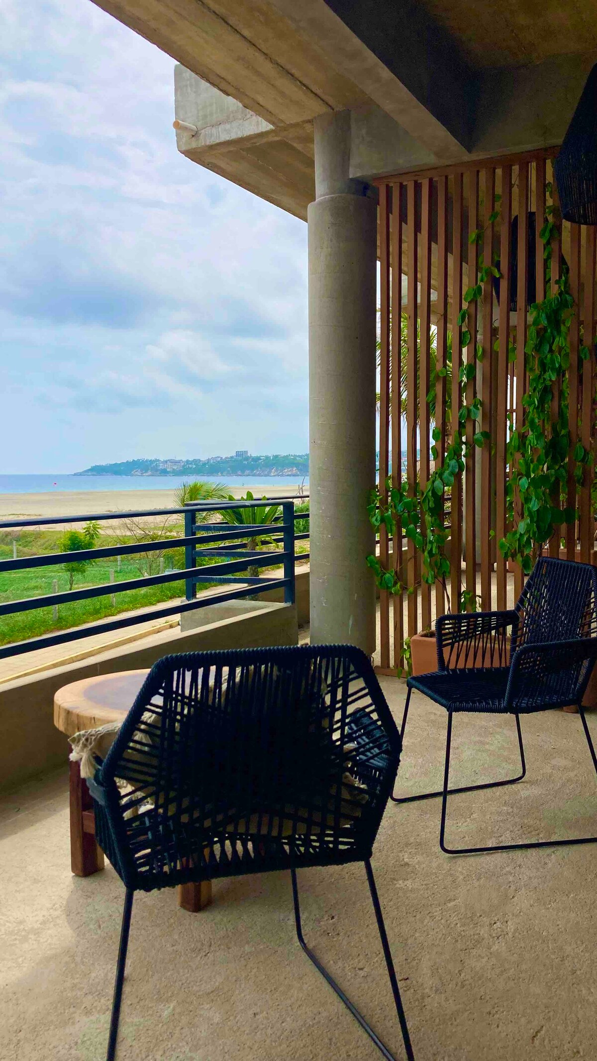 An expansive balcony offers a view of the ocean and nearby beach. Two black chairs are positioned around a round wooden table. Lush greenery climbs the vertical wooden slats, adding a touch of nature to the space. The scene is framed by a soft sky and a distant shoreline.