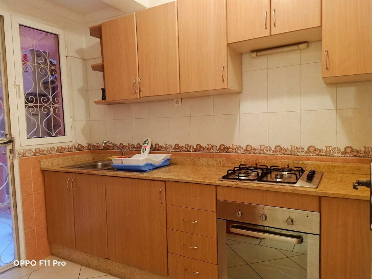 The kitchen features wooden cabinetry, a stainless steel gas stove, and a double sink. A countertop runs along one side, complementing the tiled back wall. Natural light enters through a small window above the sink, enhancing the functional layout of the space.
