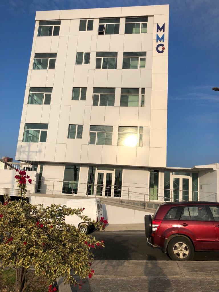 A modern, multi-story building features a sleek white façade accented with large glass windows. The entrance is framed by a gentle slope of concrete with accessible pathways. A red vehicle is parked in the foreground, while vibrant flowering plants enhance the surroundings.
