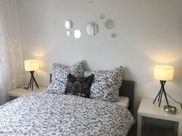 A spacious bed is centered against a neutral wall, adorned with a patterned duvet and decorative pillows. Bedside tables with modern lamps are positioned on either side, providing soft lighting. A series of decorative mirrors are mounted above the bed, enhancing the room's depth.
