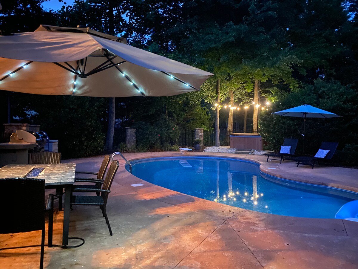 An illuminated backyard scene is set around a private pool, showcasing a patio with a dining table under an umbrella. Soft lighting from string lights and adjacent trees creates a relaxing ambiance, while a hot tub and lounge chairs are visible nearby.