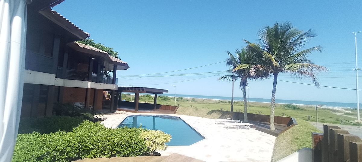 The image showcases a well-maintained outdoor area featuring a sparkling pool surrounded by lush greenery. In the background, the ocean can be seen under a clear blue sky, with a modern two-story house nearby complemented by palm trees.