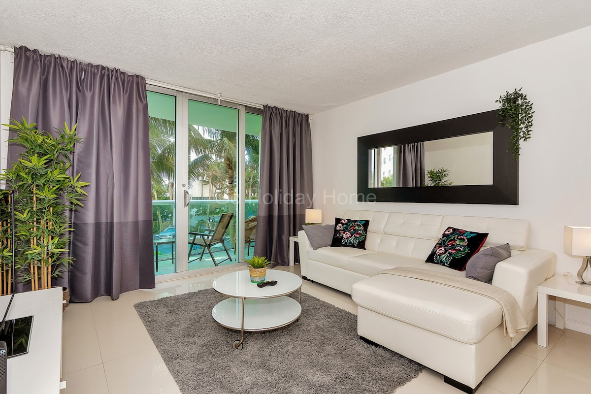 A light-filled living area features a white sectional sofa and a round coffee table on a soft gray rug. Large sliding glass doors are draped with dark curtains, revealing glimpses of outdoor greenery. A mirror and decorative plants enhance the space's modern aesthetic.
