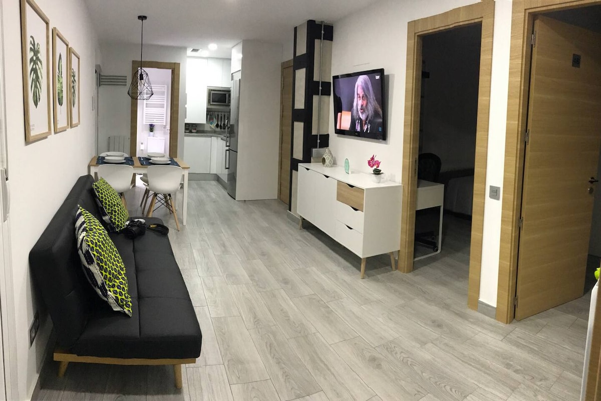 A modern living space is highlighted with a sleek black sofa adorned with decorative cushions. A dining table sits nearby, accompanied by stylish chairs. A wall-mounted TV displays a muted image. Light wood flooring complements the bright walls, creating a welcoming ambiance.