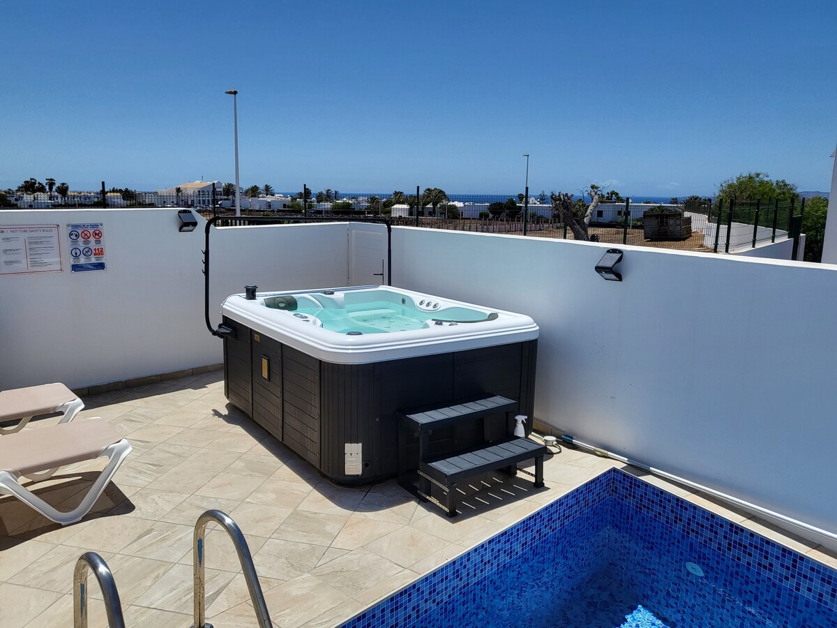 A private terrace is visible, featuring a spacious jacuzzi beside a small pool. The jacuzzi's seating accommodates three. Light tiles cover the ground, while a clear blue sky is overhead, with distant views of the coast and nearby residences beyond the terrace walls.