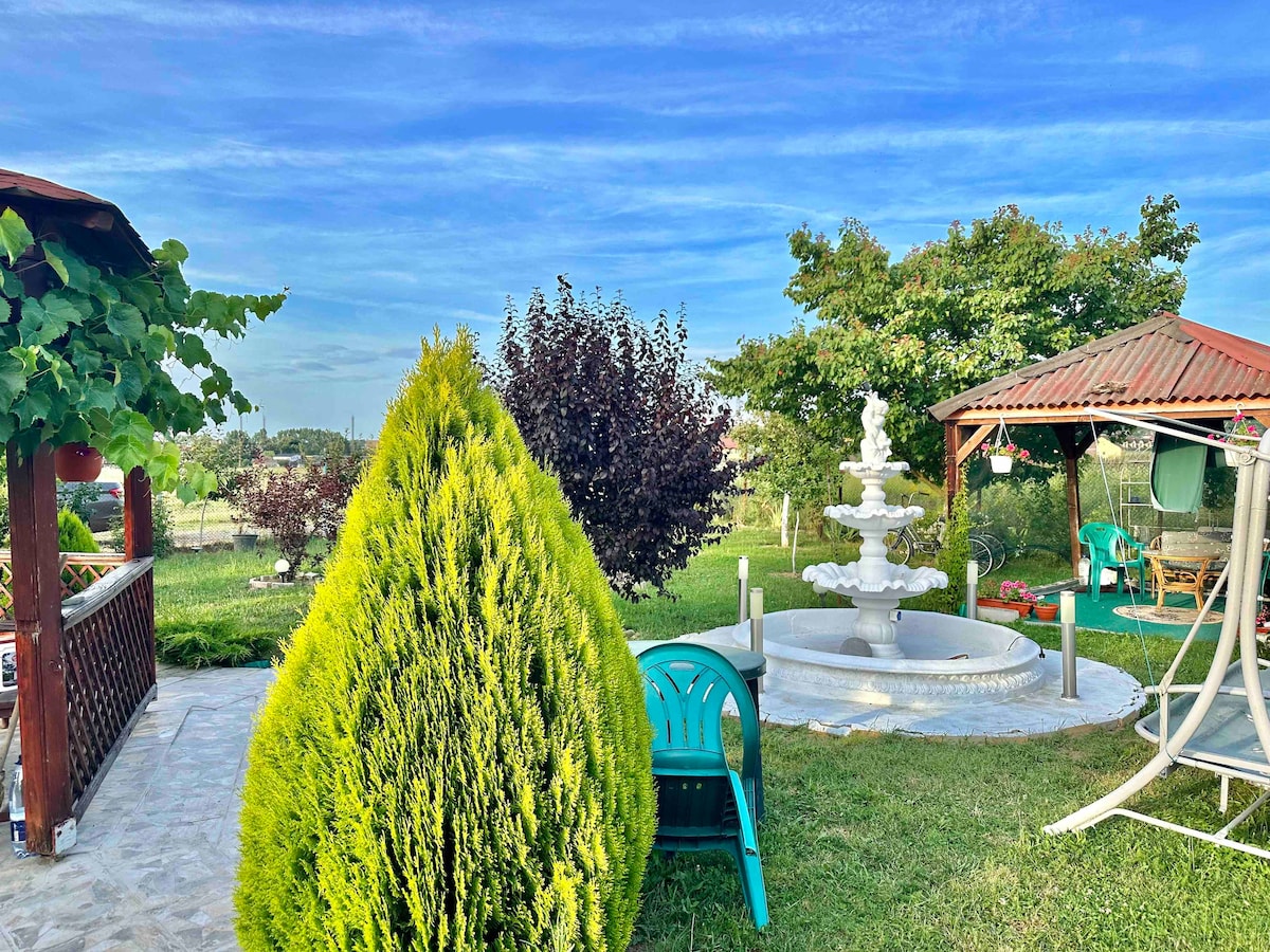 The outdoor area features a decorative fountain surrounded by lush greenery. Two gazebos provide shaded seating options, while a vibrant hedge adds color to the space. A clear sky completes the serene atmosphere, offering a peaceful retreat for guests.