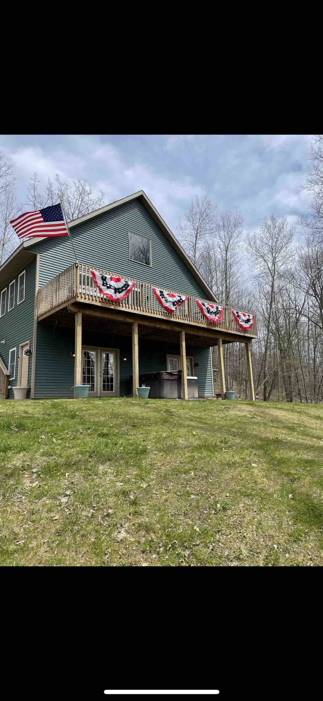 A green two-story house is situated on a grassy slope, featuring a spacious deck adorned with red, white, and blue decorations. An American flag gently waves in the breeze. Tall trees in the background portray the surrounding forested area.