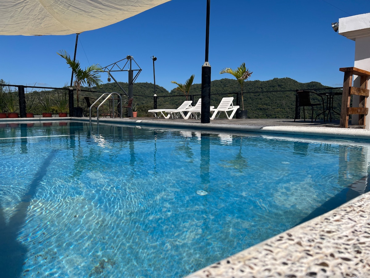 A clear swimming pool reflects the blue sky, surrounded by a sunlit terrace. Lounge chairs are positioned nearby, and lush greenery can be seen in the background, enhancing the serene setting.
