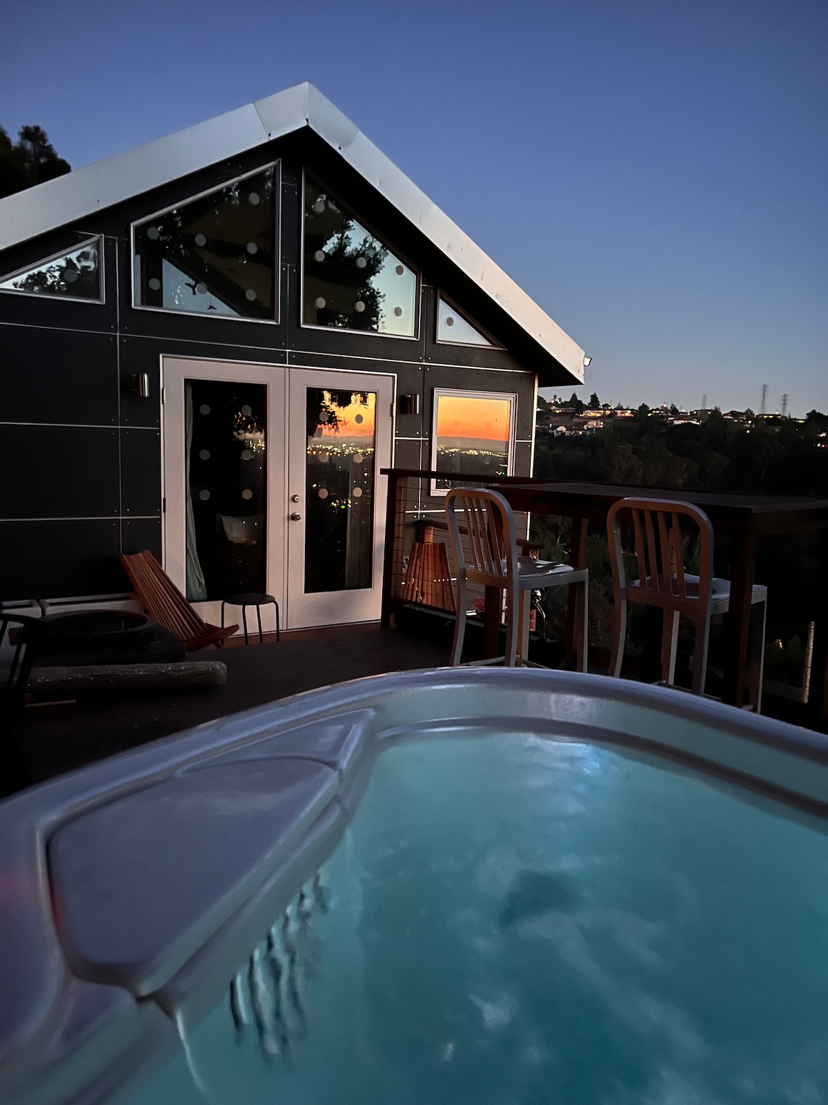 The image captures a modern cabin at dusk, showcasing large glass doors reflecting the colorful sunset. A hot tub is in the foreground, surrounded by wooden deck furniture, highlighting the tranquil outdoor space amidst a serene hillside setting.