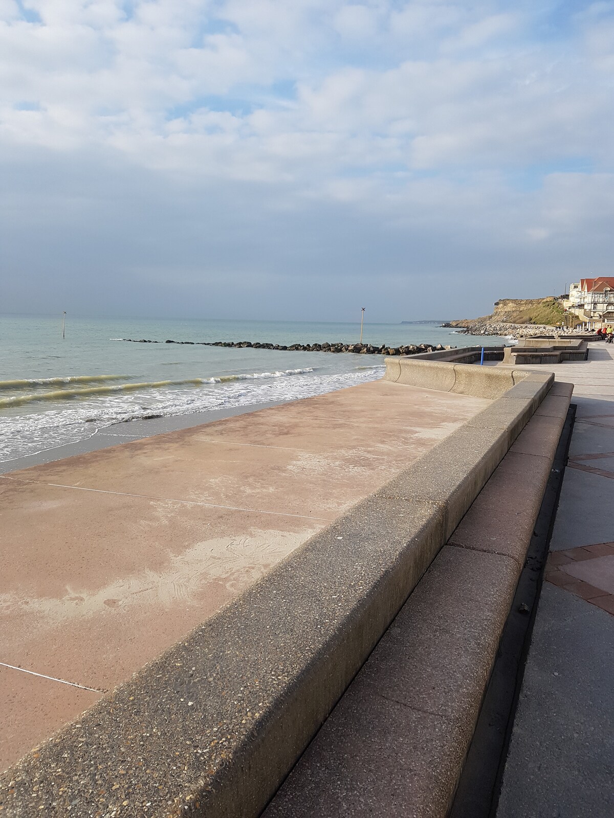 A view of a peaceful promenade along the shoreline, featuring a smooth concrete surface. The gentle waves lap against the beach, with small rocks visible in the water. Overcast skies create a soft light, while distant cliffs and buildings line the coast.