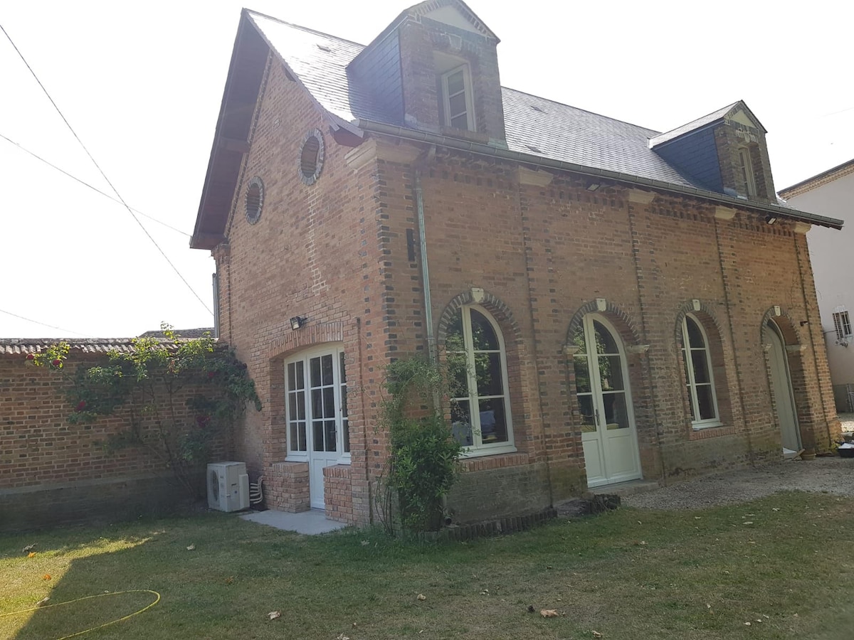 The exterior of a charming brick building is shown, featuring arched windows and a sloped roof. A grassy area surrounds the structure, with a small air conditioning unit visible on one side. The overall setting is tranquil, reflecting the quaint nature of the surrounding village.