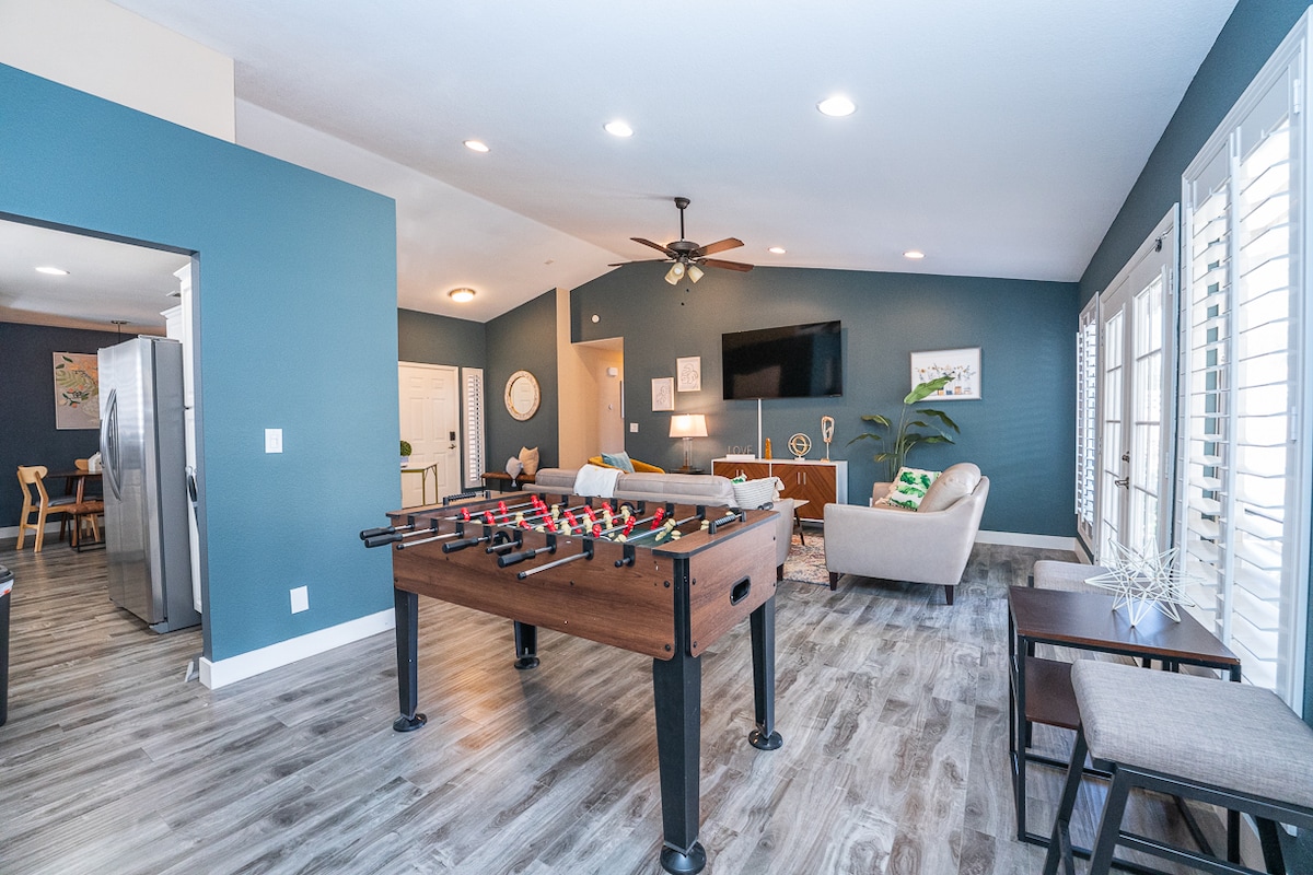 A spacious common area features a foosball table prominently in the center. The room is adorned with light-colored wood flooring and walls painted in a calming blue shade. Comfortable seating options are arranged around a mounted television, with natural light filtering through multiple windows.