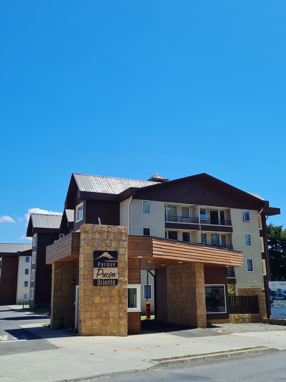 A modern multi-story building is depicted, featuring a mix of rustic and contemporary architectural elements. The entrance includes a stone structure with the name 'Parque Pucón' prominently displayed. Landscaping and blue skies enhance the welcoming appearance of this central location.