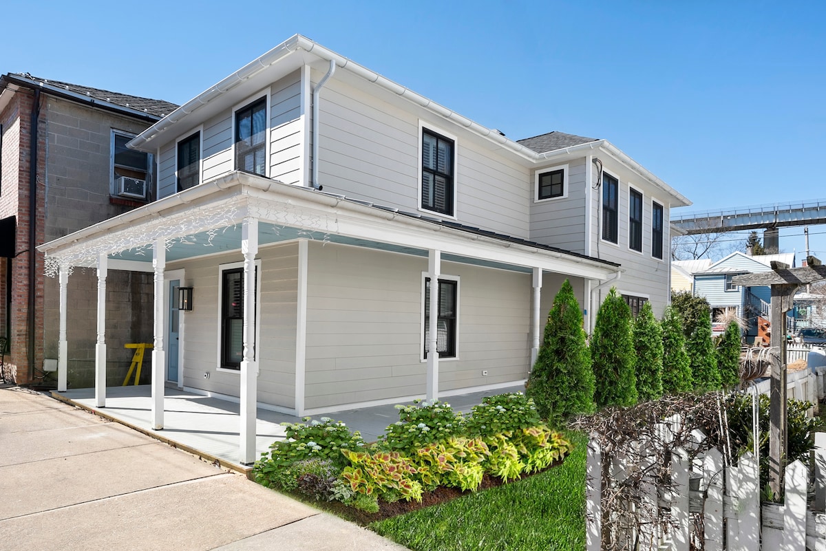 A newly renovated two-story house features a large front porch with white columns, offering ample space for relaxation. Decorative trim adds charm, while neatly arranged greenery and low shrubs border the yard. The driveway provides two off-street parking spaces beside a well-maintained path.