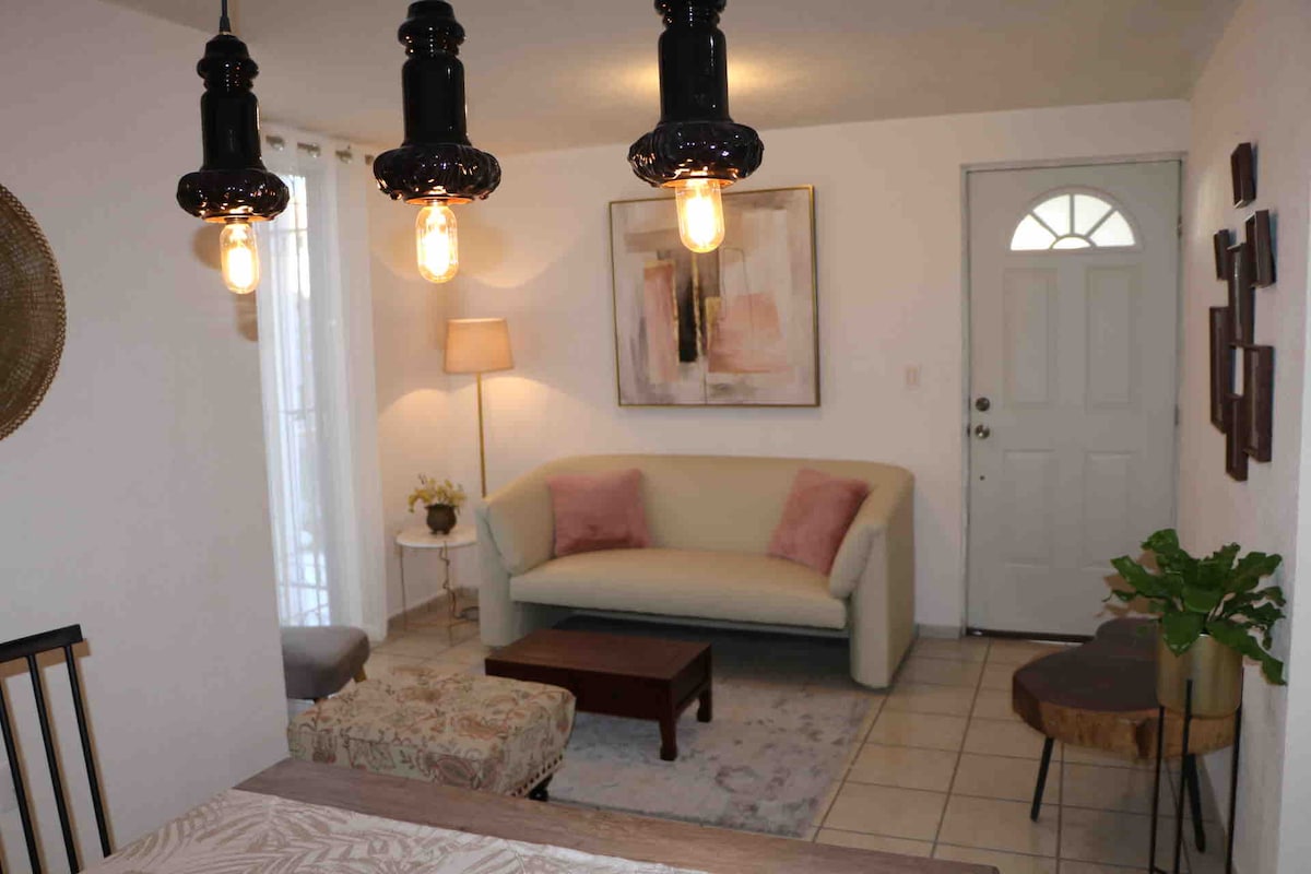 A bright living area features a light-colored sofa with soft cushions and a small coffee table. Pendant lights hang above, illuminating the space. A door leads outside, and a decorative plant is placed beside a side table, enhancing the room’s inviting ambiance.