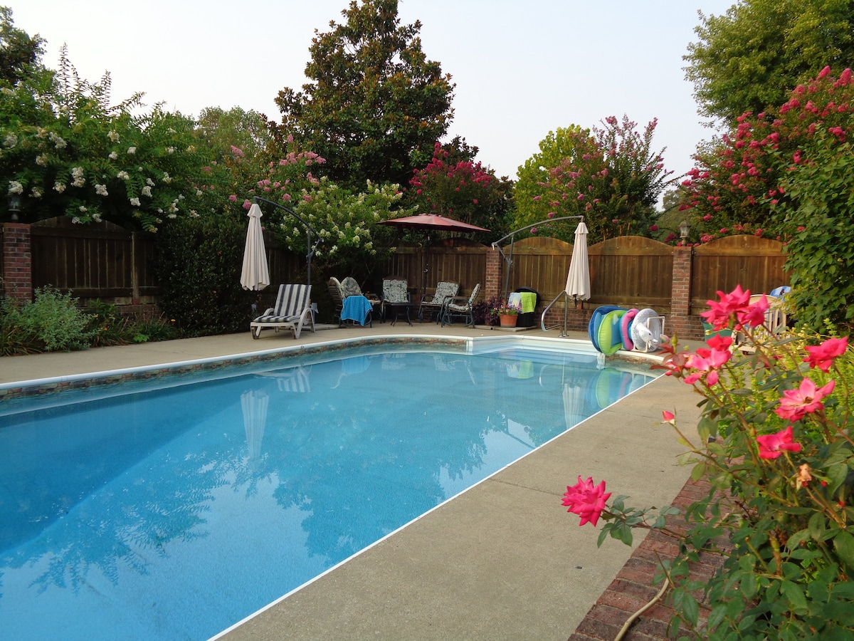 An inviting in-ground pool is surrounded by lush greenery and blooming flower bushes, creating a serene outdoor space. Lounge chairs are arranged nearby, with additional seating available under a shady patio. Colorful pool floats rest along the pool edge, enhancing the relaxing atmosphere.