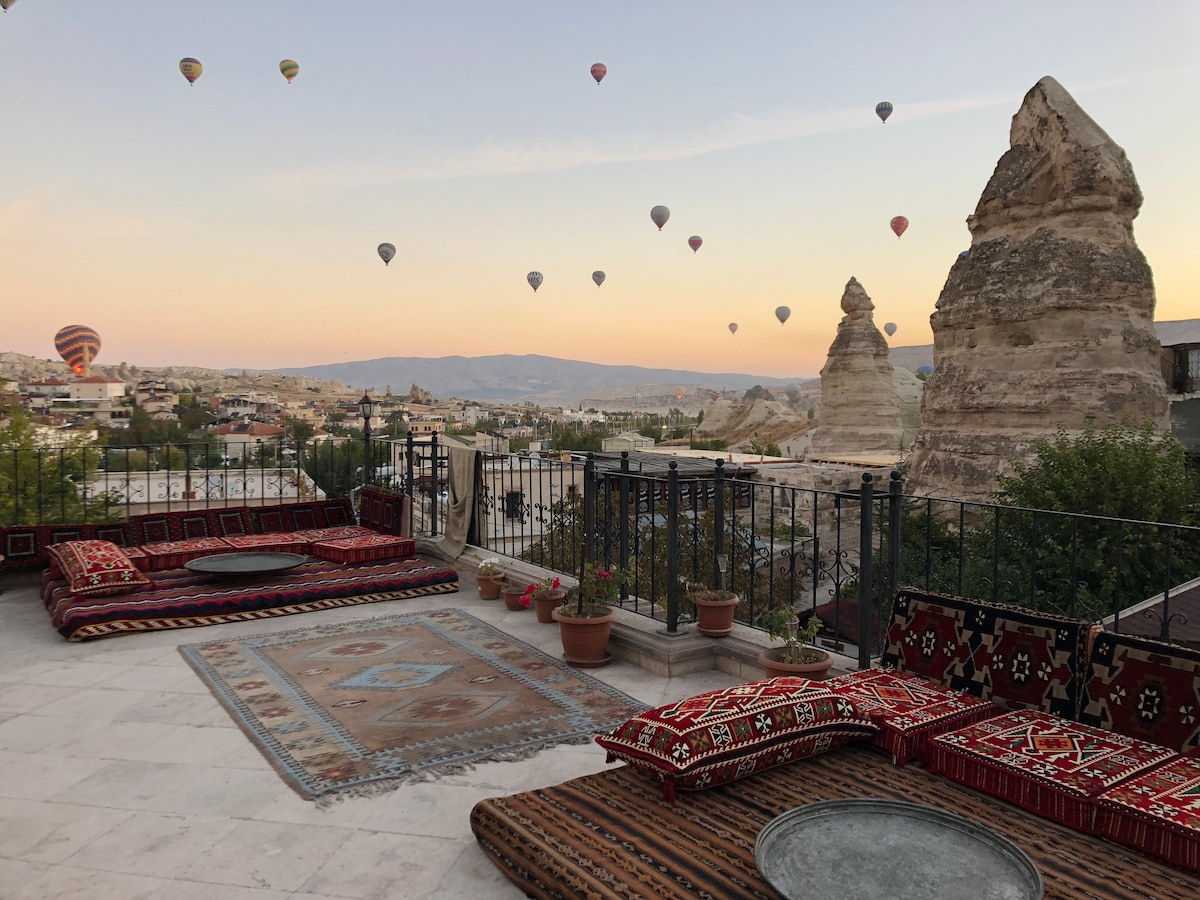 A spacious terrace features traditional seating with colorful cushions and textiles, complemented by a central low table. The backdrop showcases multiple hot air balloons floating above the scenic landscape, with fairy chimney rock formations visible in the distance against a soft sunset sky.