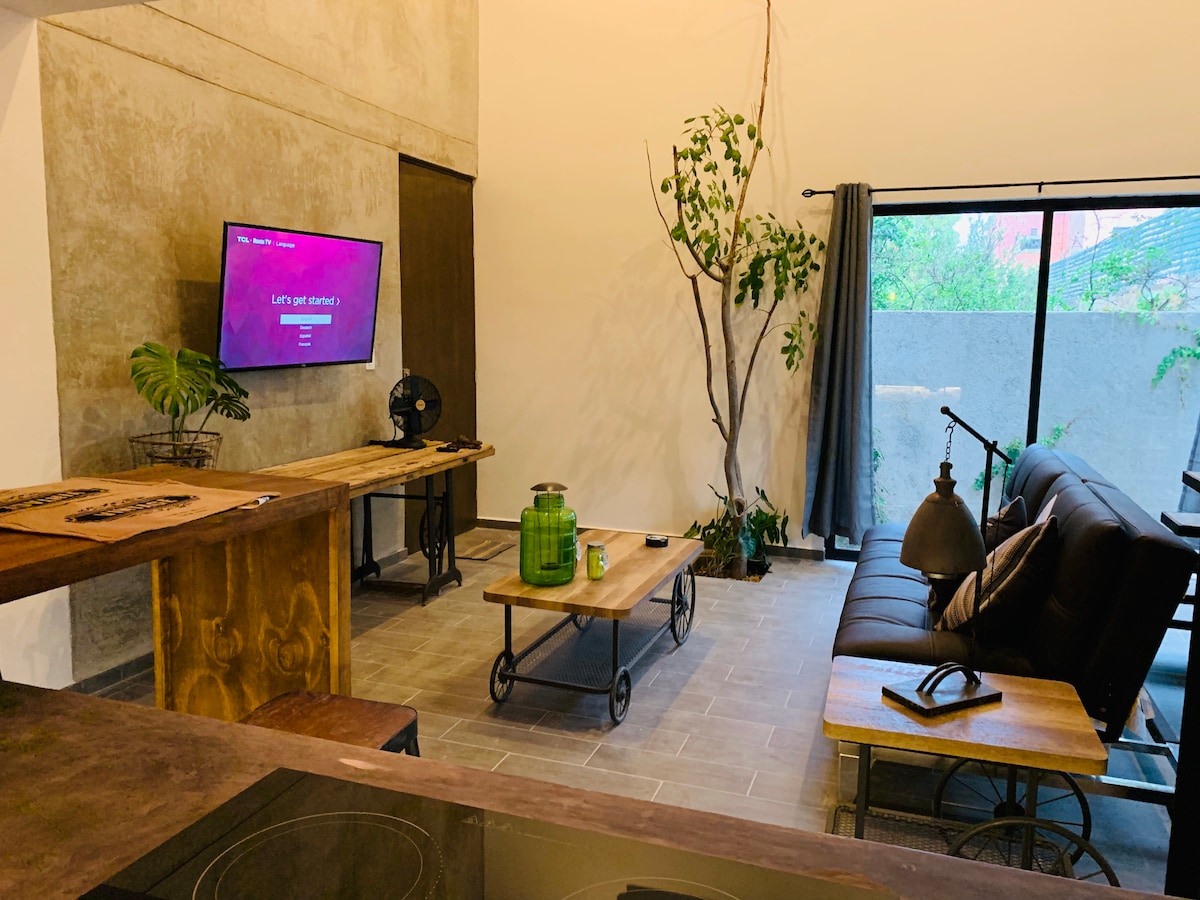 A sunlit living area features a comfortable seating arrangement and a wall-mounted smart TV. A wooden breakfast bar complements rustic decor elements, with plants adding a touch of greenery. Large glass doors open to an outdoor space, enhancing the overall lightness of the room.