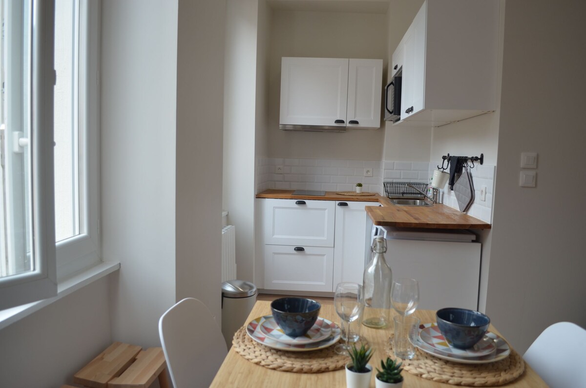 The kitchen area features white cabinetry and wooden countertops. A dining table is set with dishware and glassware. Sunlight enters through a window, highlighting the modern appliances and a small potted plant on the table, creating a functional and welcoming space.
