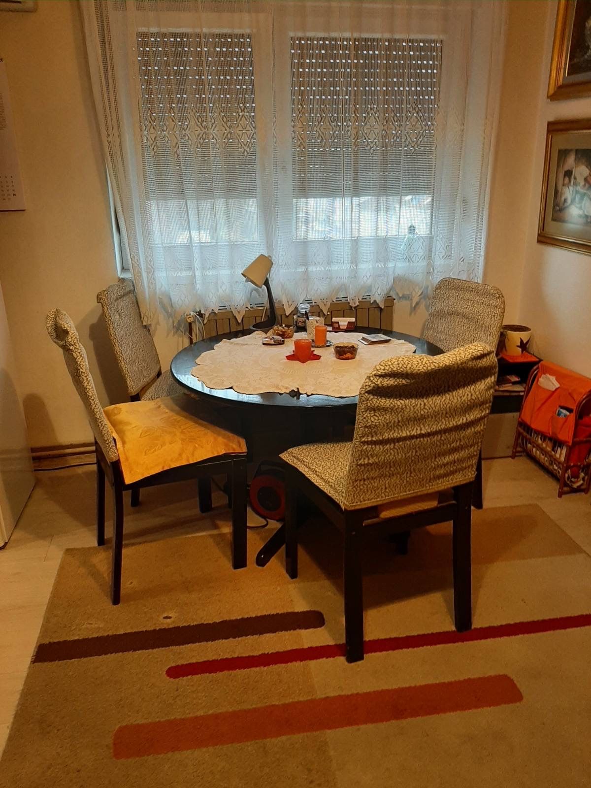 A round dining table is surrounded by four chairs, featuring woven fabric cushions. A light-colored tablecloth covers the table, which displays various decorative items. Natural light enters through curtained windows, while a patterned rug anchors the dining area.