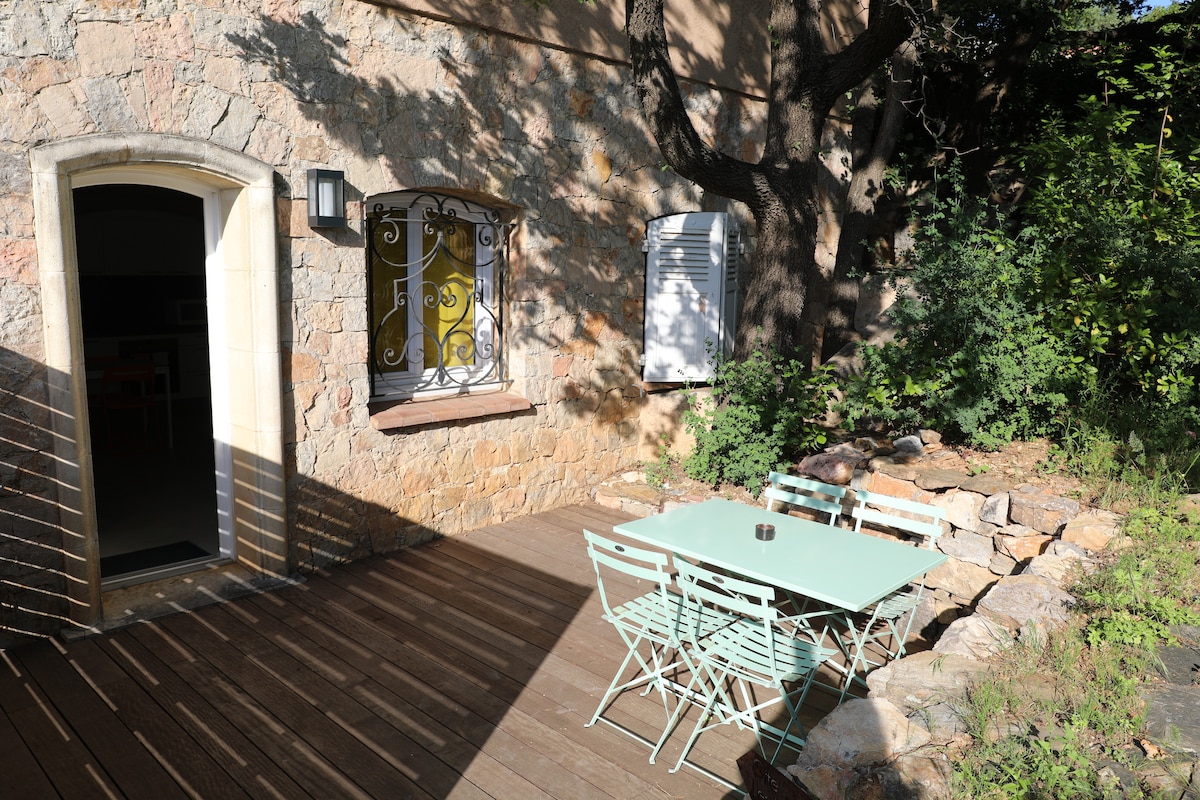 An outdoor seating area features a light green table surrounded by matching chairs on a wooden deck. The space is framed by a stone wall and shaded by trees, with a door leading into the interior of the villa.