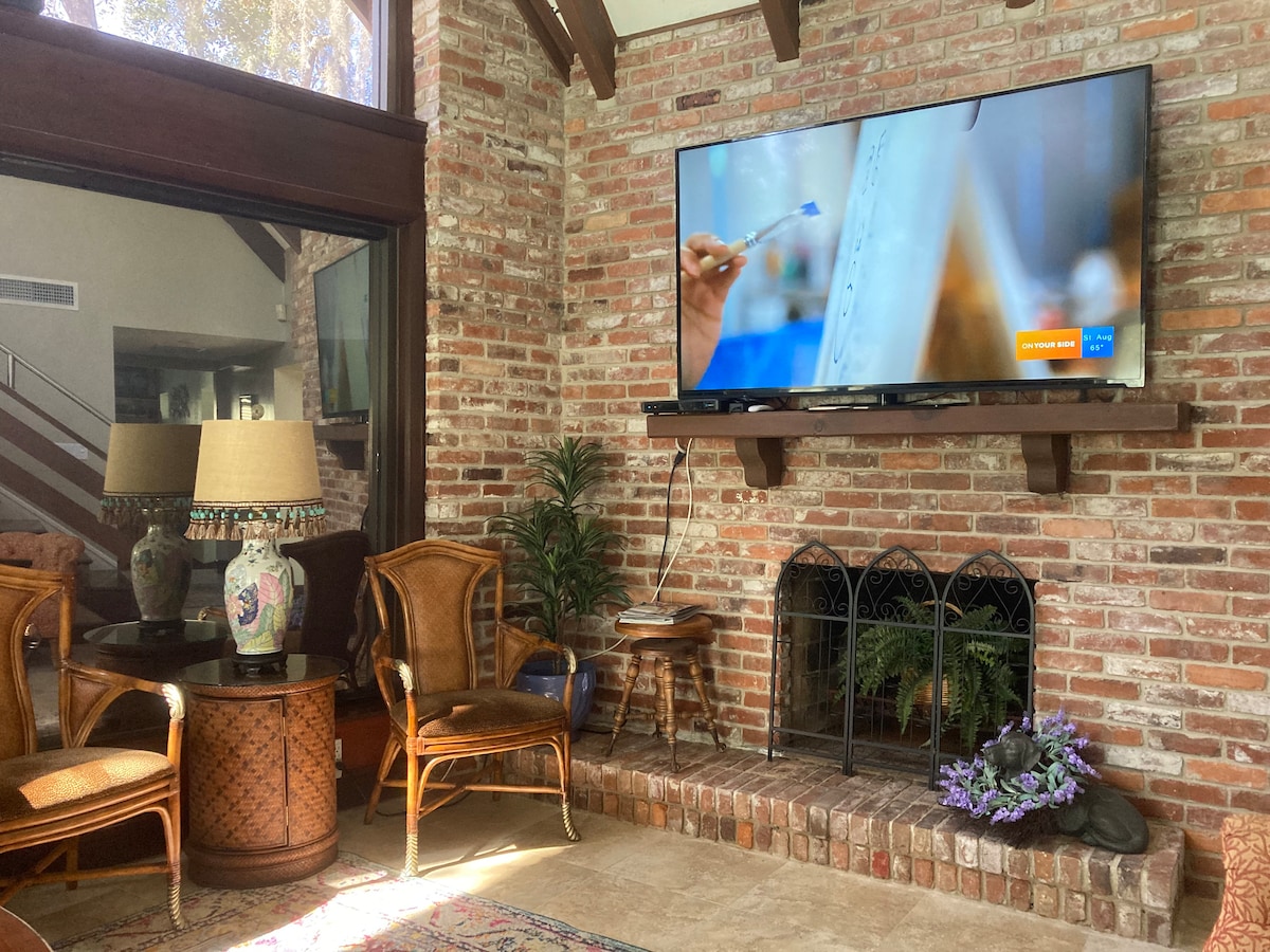 A cozy living area showcases a mounted television above a brick fireplace. Two stylish rattan chairs are positioned beside a round table, which features a decorative lamp. Natural light filters through large windows, illuminating the space with a warm glow.