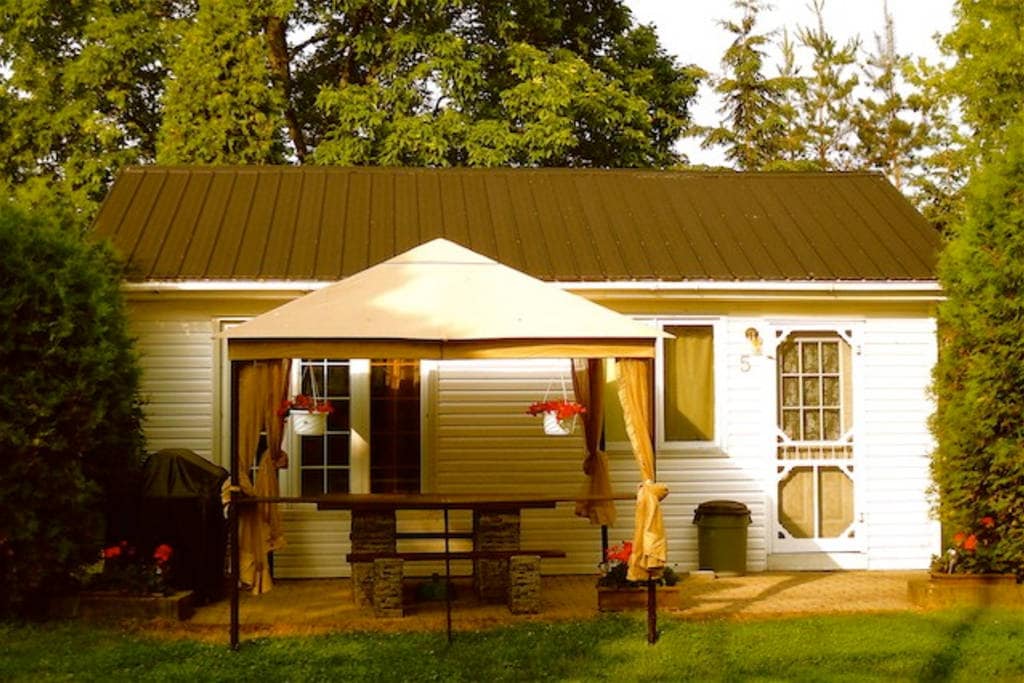The exterior of a one-bedroom cottage is showcased, featuring a light-colored façade and a welcoming entrance. A large gazebo with flowing curtains provides a shaded area over a picnic table, surrounded by greenery and flowerpots, enhancing the outdoor space.