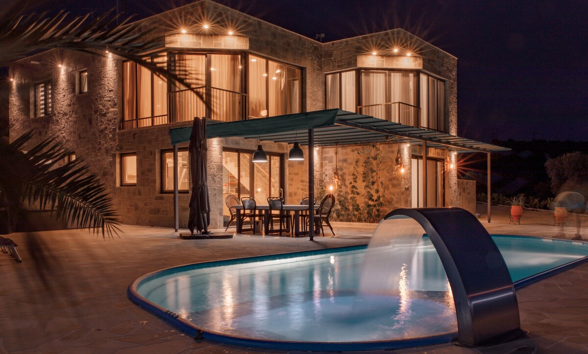 The villa's exterior is illuminated at night, highlighting a stylish pool with a waterfall feature. An outdoor dining area is visible under a covered patio, surrounded by large glass windows that reflect warm light from within the structure.