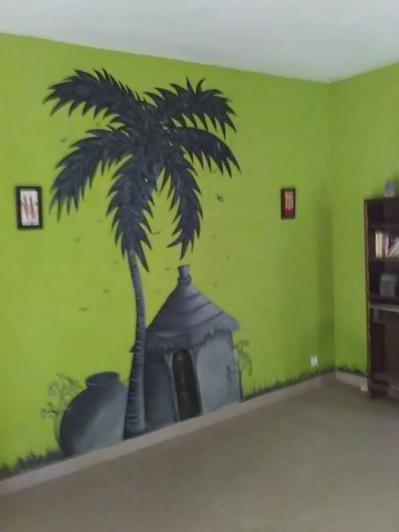 The room features vibrant green walls adorned with a mural depicting a palm tree and a traditional house. Two framed artworks are displayed on the walls, while a dark wooden shelving unit is positioned against one side, showcasing a selection of books.