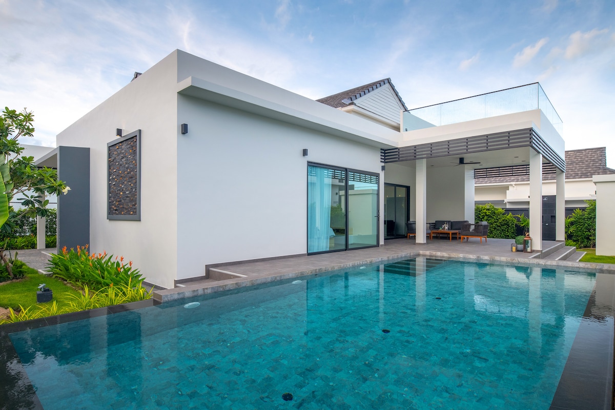 A modern villa features clean lines and large glass doors leading to a private pool. The outdoor area includes a lounge space beside the water, surrounded by well-maintained vegetation. The upper terrace is visible, offering additional outdoor seating and an open sky.