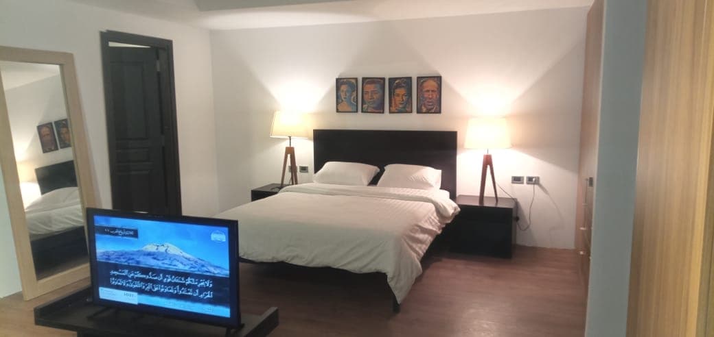 The sleeping area features a king bed framed by two bedside lamps, offering soft illumination. A sleek TV is positioned across from the bed, and a full-length mirror reflects the room's spacious design. Artwork is displayed above the bed, adding visual interest.