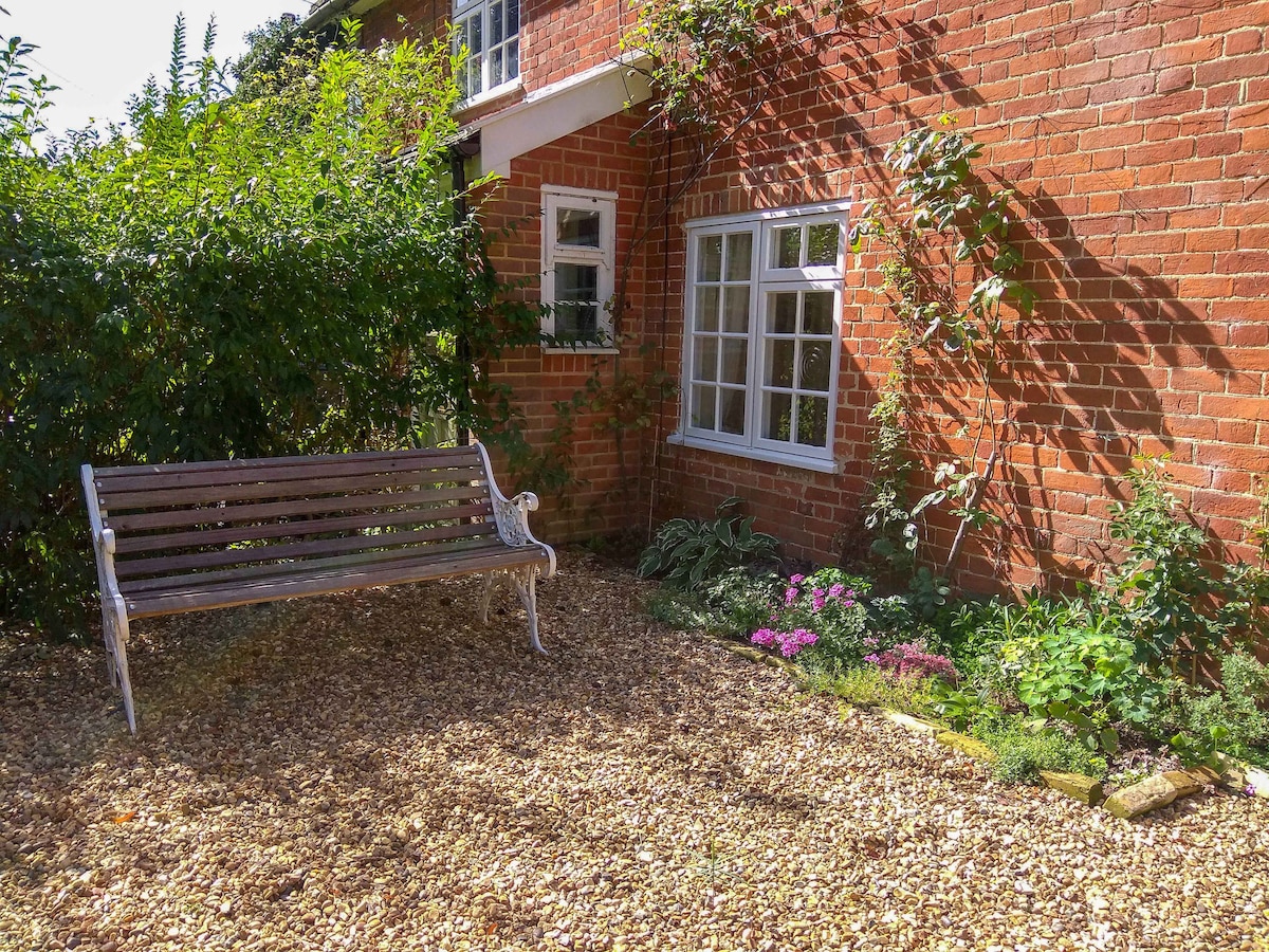 A charming outdoor area is presented, featuring a wooden bench nestled among gravel and lush greenery. Flowering plants add color, while the warm red brick wall of the cottage provides a rustic backdrop. Light filters through the foliage, enhancing the peaceful atmosphere.