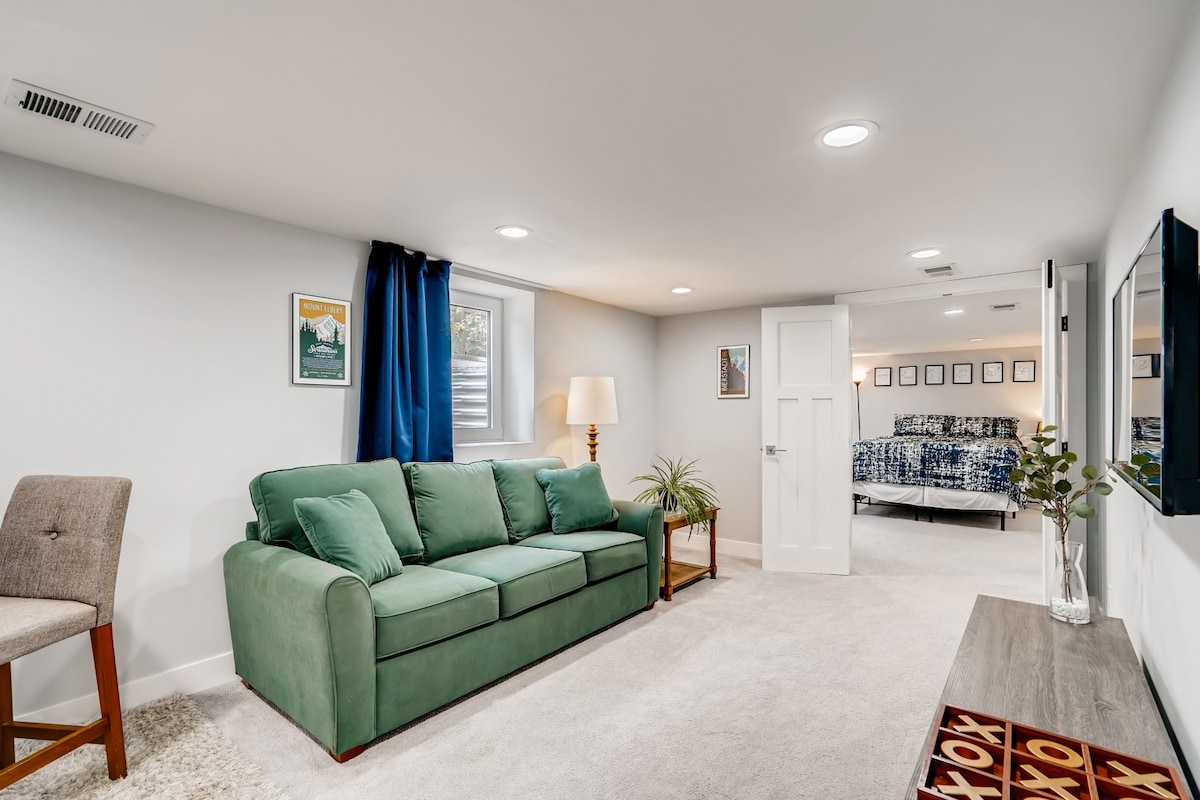 A spacious living area features a green sofa with decorative cushions, complemented by a floor lamp and a small plant. Natural light enters through a window, while a door leads to a bedroom with a neatly made bed in the background.