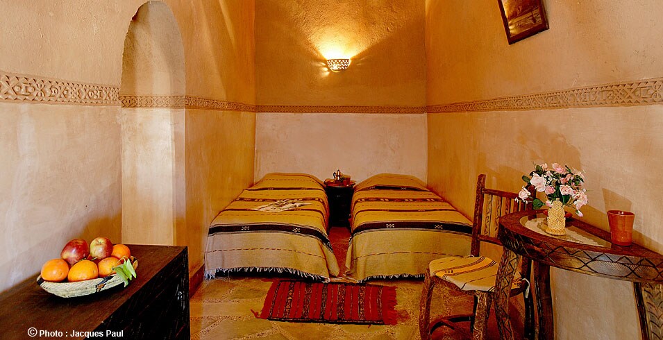 A cozy room features two single beds draped with patterned blankets in warm tones. A small wooden table holds a bowl of fruit alongside a floral arrangement. The walls are subtly textured, and a decorative border enhances the rustic charm.