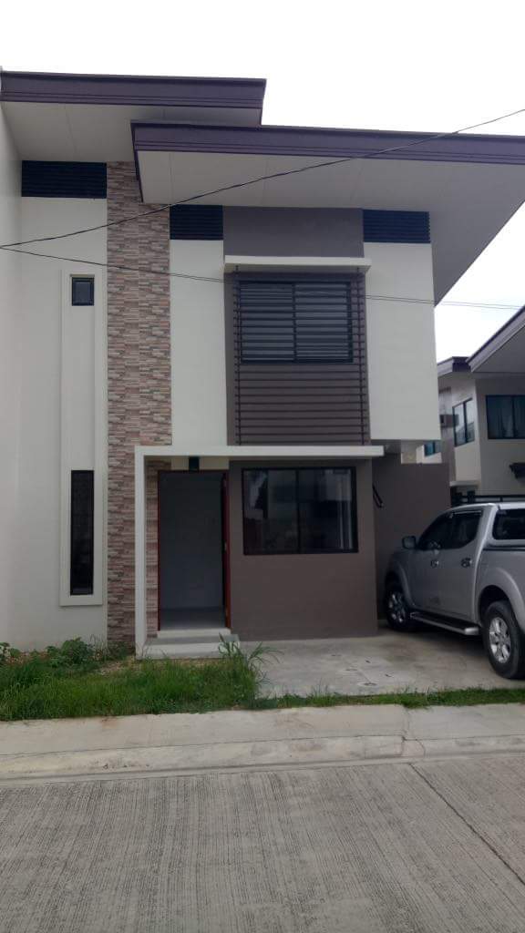 A modern two-story house features a combination of textured brick and smooth exterior walls. Large windows allow natural light into the space, while a driveway accommodates a parked vehicle. Minimal landscaping surrounds the entrance, set against a clear concrete pathway.
