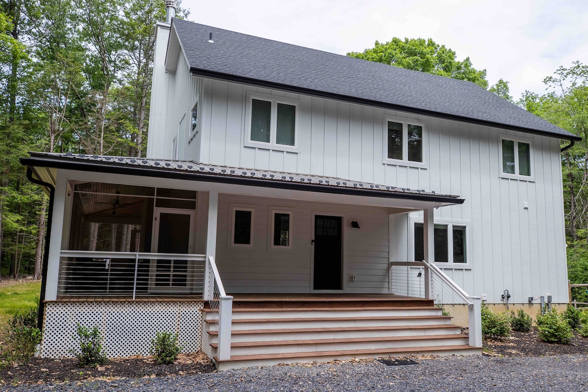 The exterior of a modern farmhouse is showcased, featuring a two-story structure with a sleek white façade. A welcoming front porch with wooden steps leads to the main entrance, which is flanked by several windows. Lush greenery surrounds the home, enhancing the natural setting.