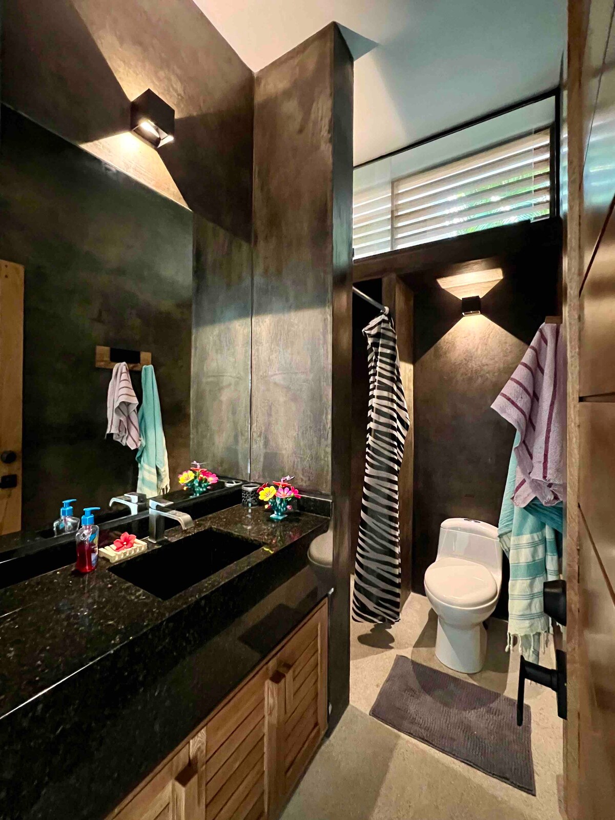 A modern bathroom is presented with dark walls and a polished black granite countertop. A white toilet is positioned behind a wooden door. Colorful flowers and towels are neatly arranged, contributing to the aesthetic. Soft lighting highlights the space, creating a functional and stylish atmosphere.
