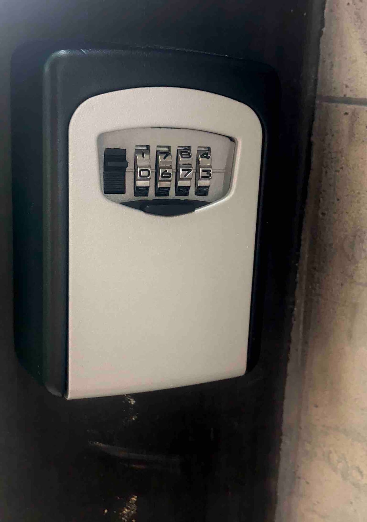 A secure lockbox is mounted on a wall, featuring a combination keypad. The box is designed for key storage, with a neutral color scheme and a sturdy appearance, positioned in a well-lit area that ensures visibility.