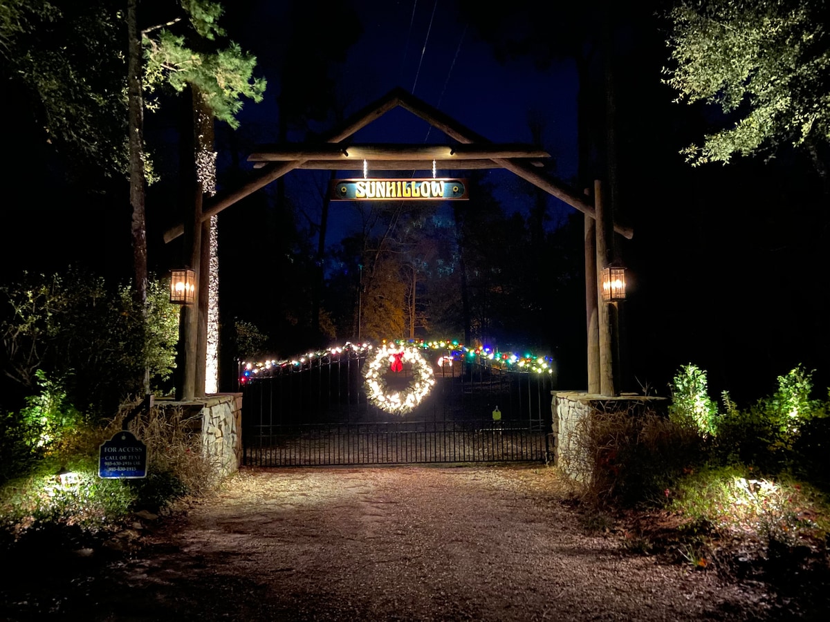 An illuminated entrance gate to the property is adorned with festive lights and a wreath. The surrounding trees provide a natural backdrop, while the gravel pathway leads inward, creating an inviting entryway to the secluded retreat.