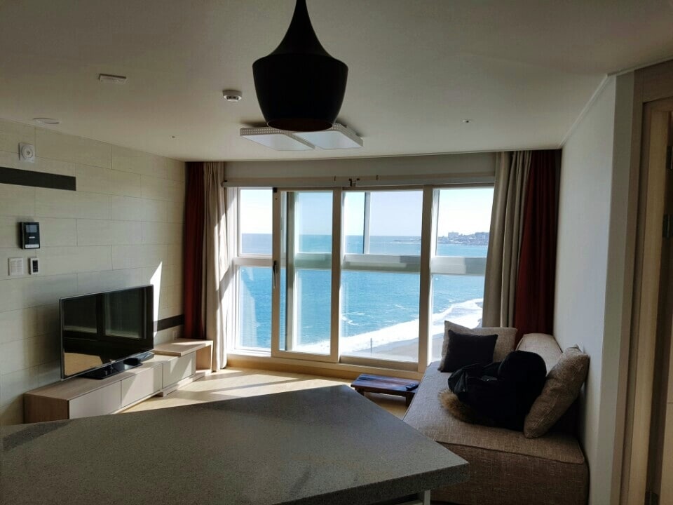 A light-filled living room features large windows that offer an expansive view of the ocean. A flat-screen TV is positioned across from a comfortable sofa, while a dining area with a stone countertop is visible in the foreground.